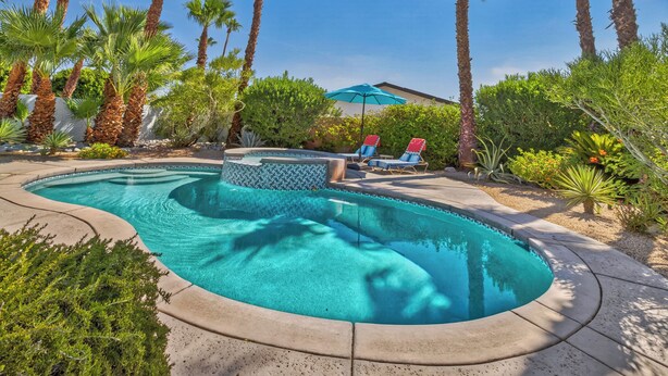 A swimming pool with two lounge chairs and a blue umbrella, surrounded by palm trees and greenery.