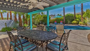 An outdoor patio with a covered seating area, a pool, and surrounding greenery.