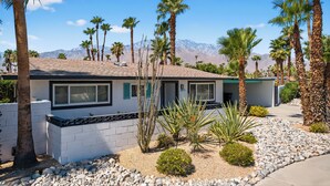 A modern two-story house with a stone foundation, surrounded by a desert landscape with palm trees and mountains in the background.