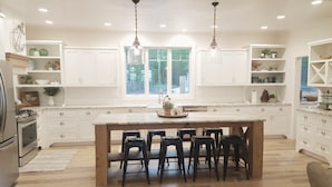 A spacious kitchen with white cabinetry, a large island with bar stools, pendant lighting, and a view of the outdoors through the window. - North Pine Cottage #1 (Brainerd)