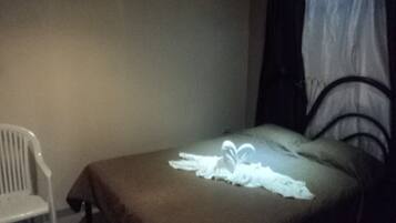 A single bed with a brown bedspread and white pillows, a white chair, and a window with a black frame and white curtains.