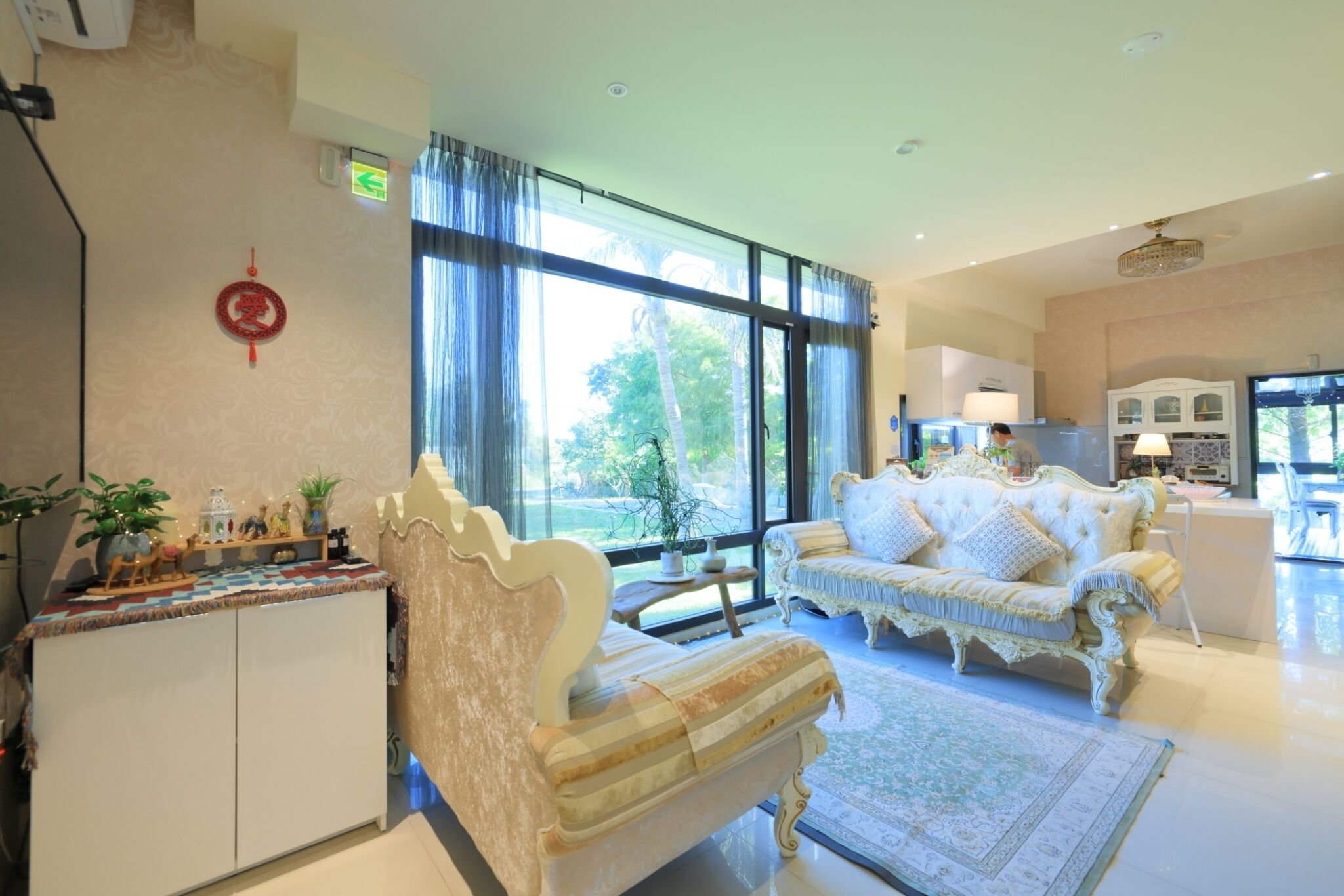 Un salón espacioso con un gran ventanal, un sofá blanco, un tapete estampado y un mueble con adornos.