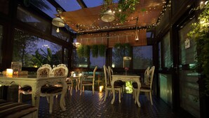 Un espace extérieur aménagé pour les repas, avec des tables et des chaises, des guirlandes lumineuses, des plantes suspendues et un sol carrelé.