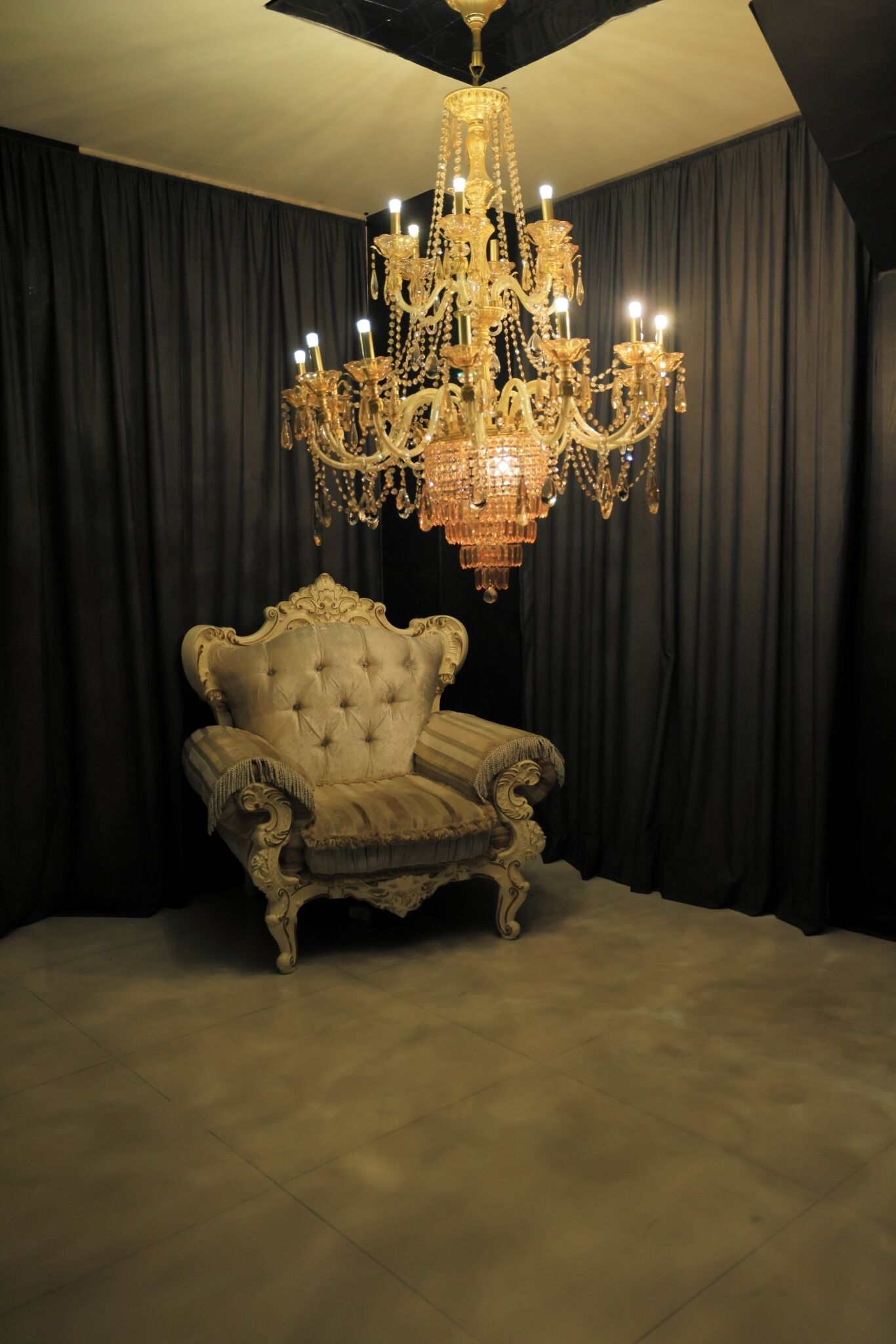 A room with a large ornate chandelier, a tufted armchair, and dark curtains.