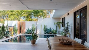 Un área de piscina moderna al aire libre con una mesa y sillas de madera, rodeada de plantas y cactus variados.