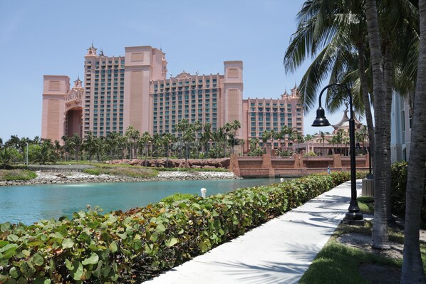 A large hotel with multiple towers, a waterfront, and a pathway lined with greenery.