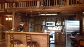 A spacious kitchen with wooden cabinetry, a central island with seating, stainless steel appliances, and a wooden staircase leading to an upper level.