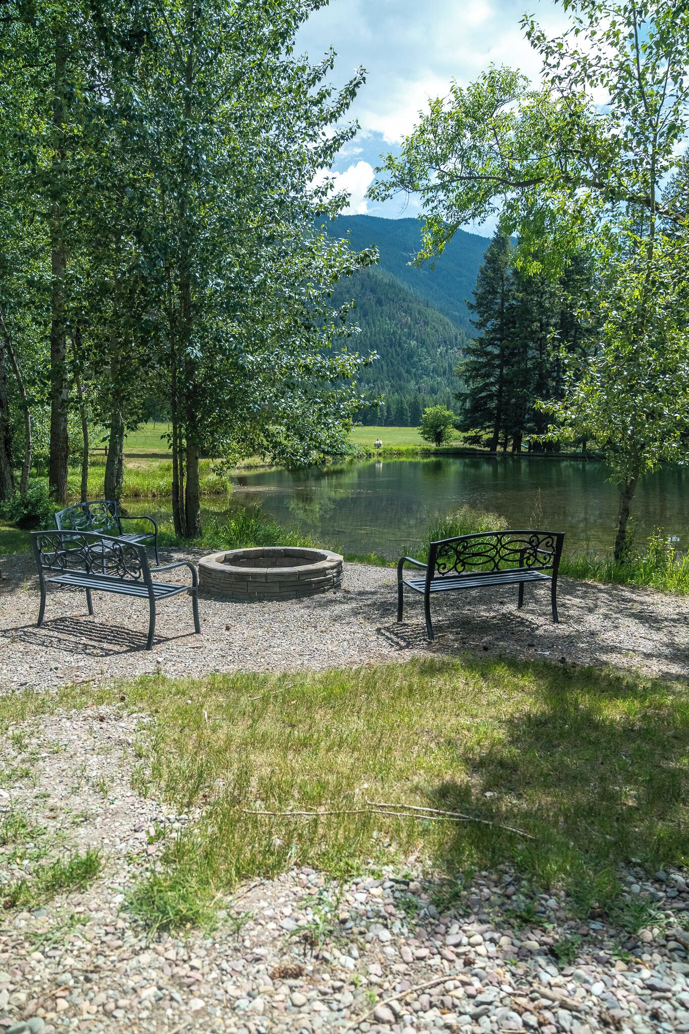 Un parc paisible au bord d’un lac, avec deux bancs en métal et un foyer, entouré d’une végétation luxuriante et dominé par une chaîne de montagnes.