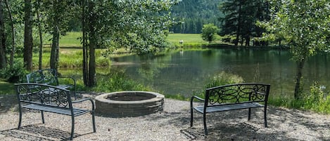Un parc paisible au bord d’un lac, avec deux bancs en métal et un foyer, entouré d’une végétation luxuriante et dominé par une chaîne de montagnes.
