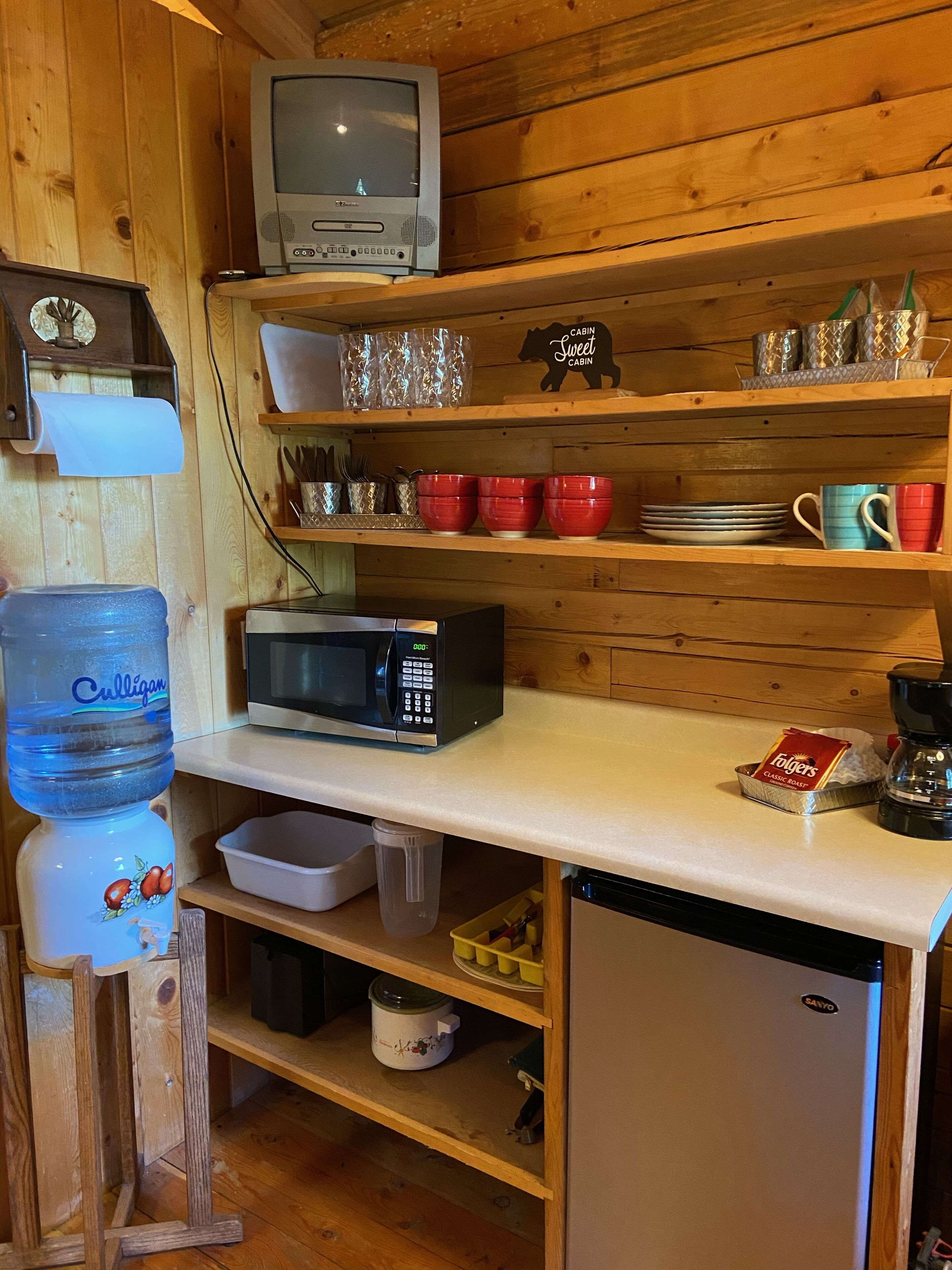 A compact kitchen with a wooden interior, a water dispenser, a microwave, a coffee maker, and a refrigerator.