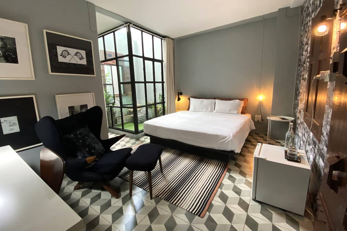 A modern hotel room with a large bed, a seating area with a chair and ottoman, a small table, and a large window with a view of greenery.