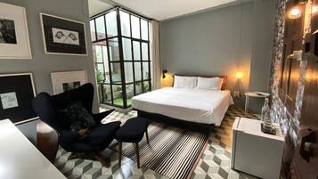 Habitación de hotel moderna con una cama grande, una zona de estar con una silla y reposapiés, una mesita pequeña y un ventanal con vistas a zonas verdes.
