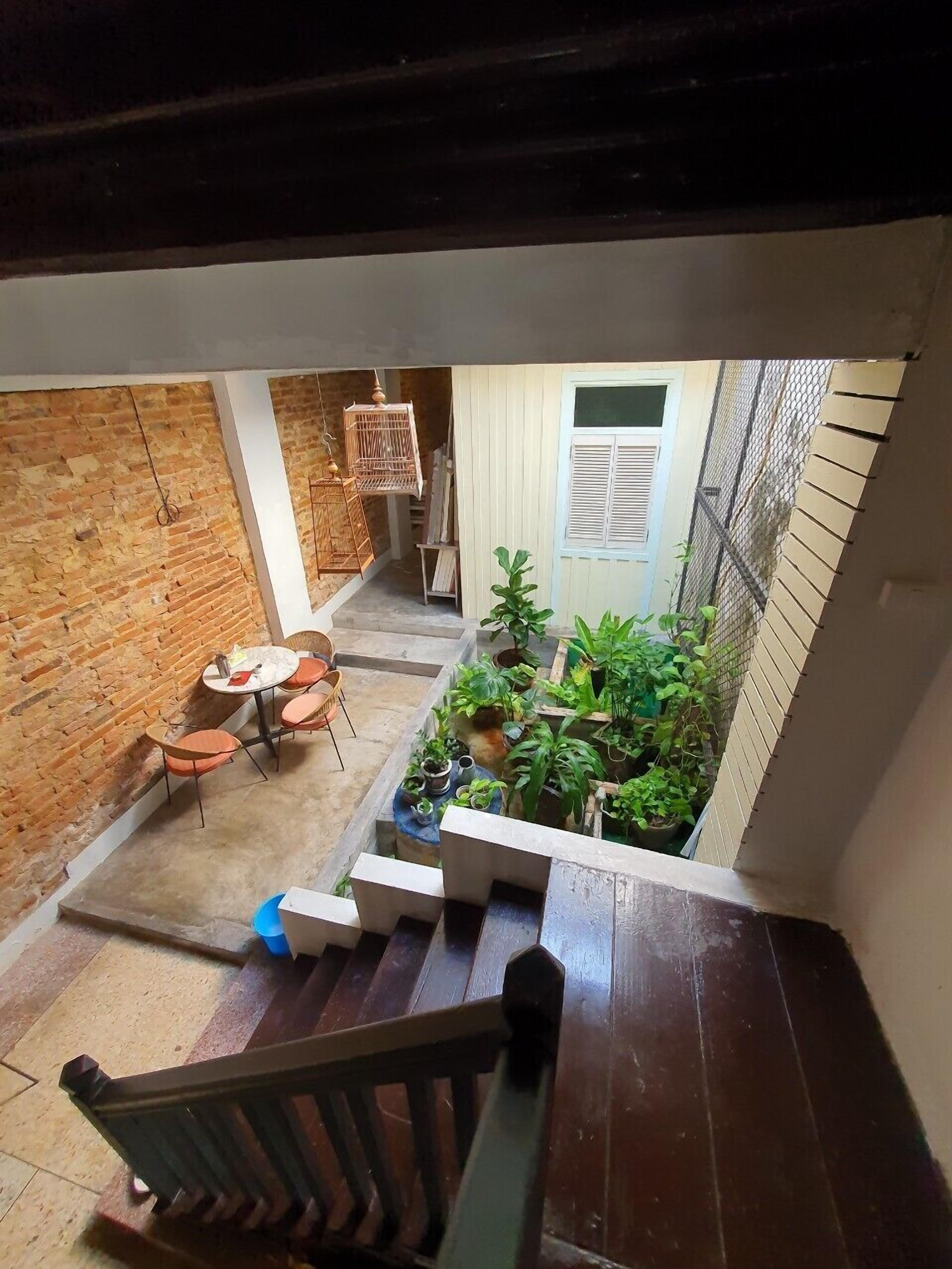 A view from a balcony overlooking a small courtyard with a brick wall, a wooden staircase, a table with chairs, and various potted plants.