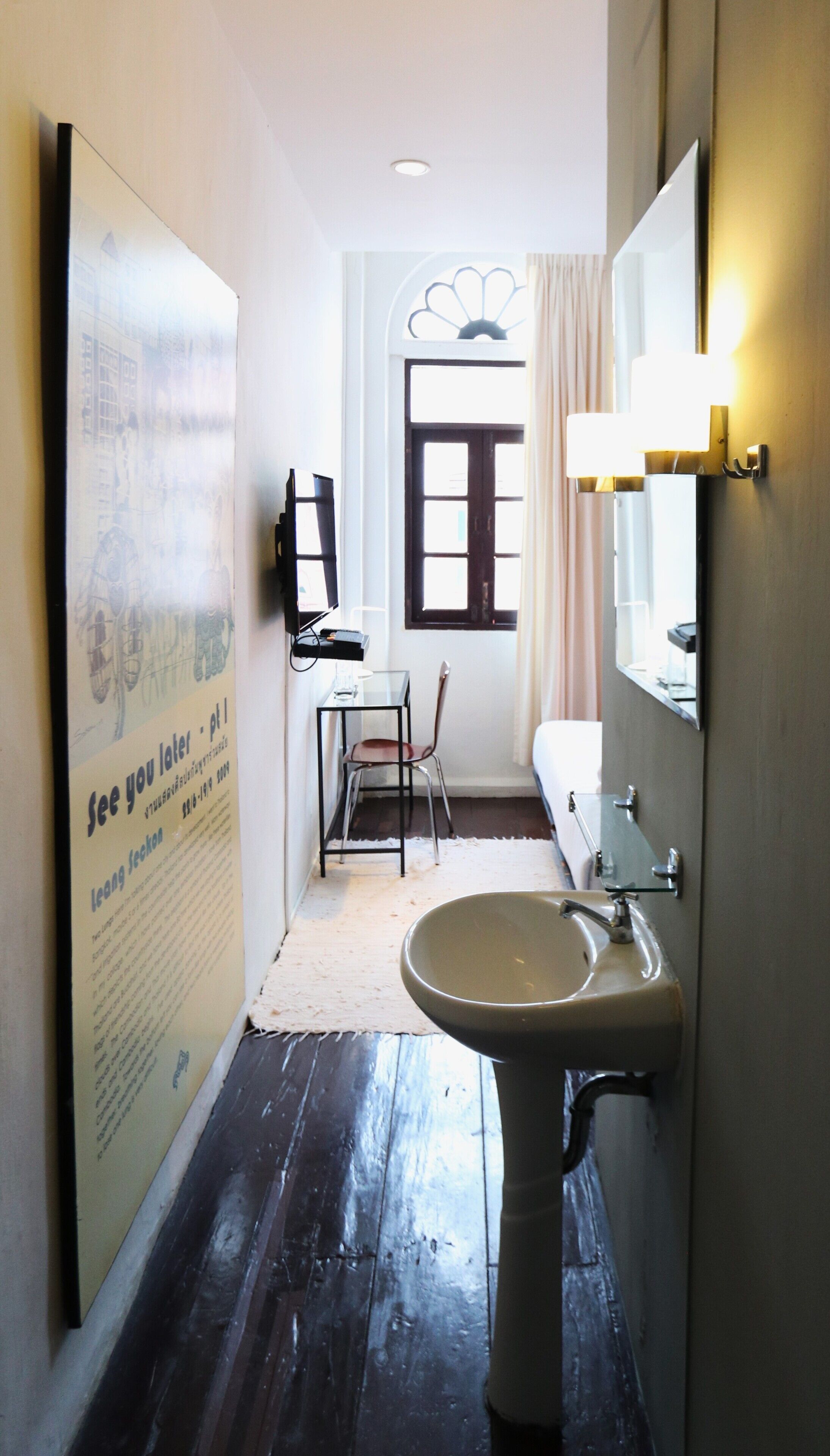 A narrow bathroom with a sink, mirror, and chair. A poster is on the wall, and a window with curtains is visible.