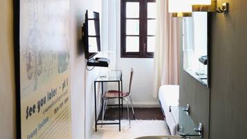A narrow bathroom with a sink, mirror, and chair. A poster is on the wall, and a window with curtains is visible.