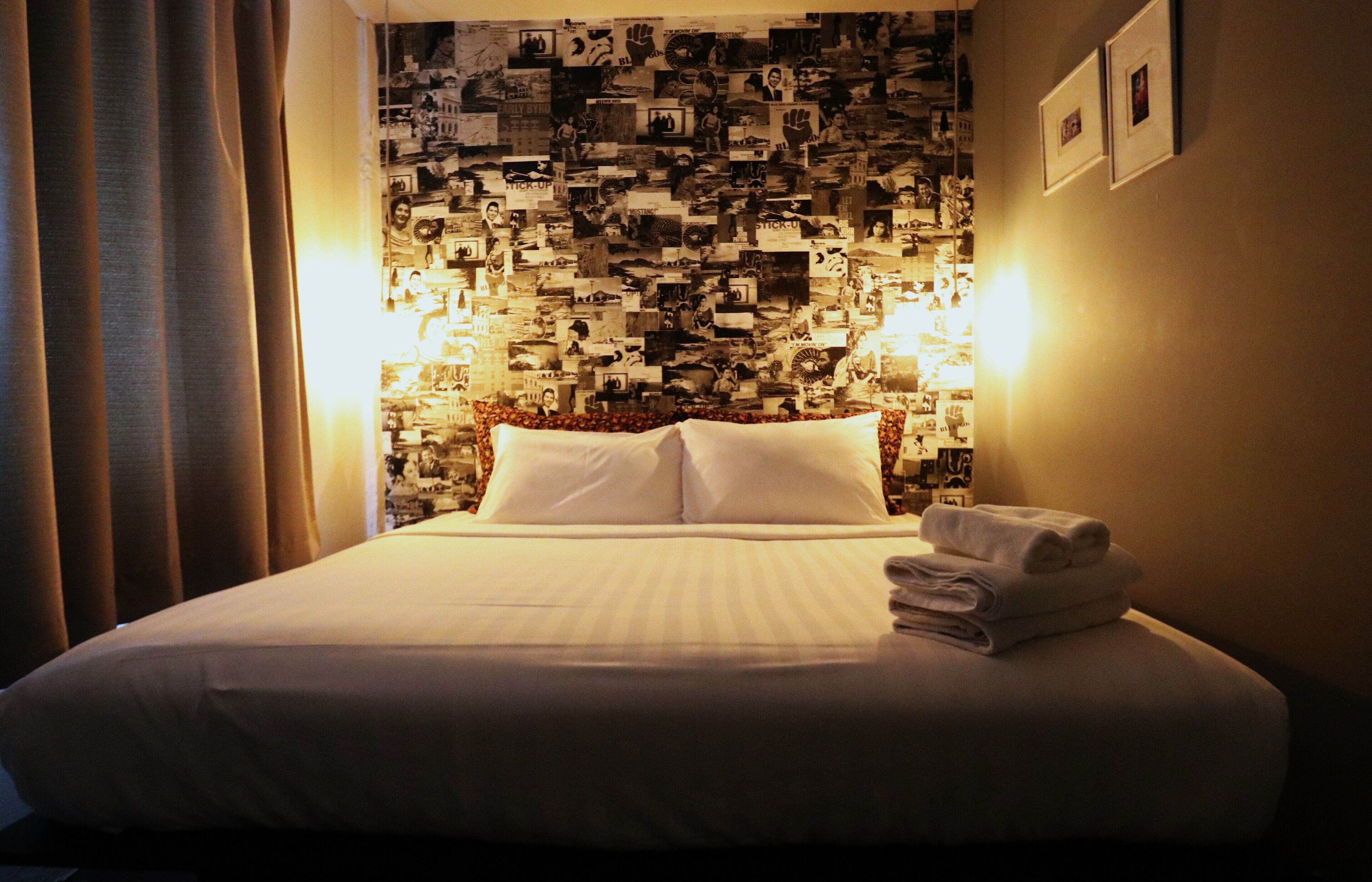 A hotel room with a bed, a wall covered with photos, and a nightstand with a lamp.