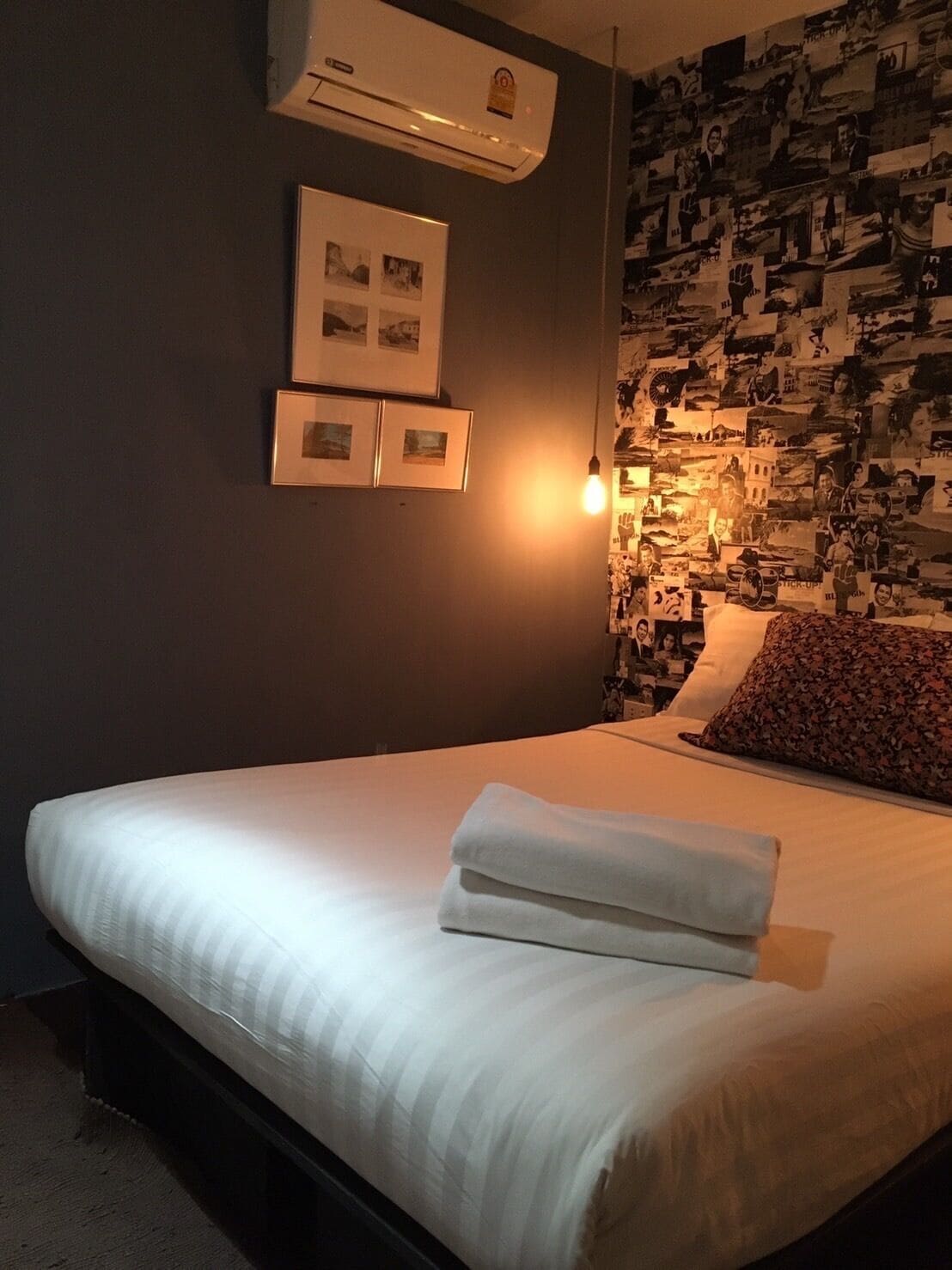 A neatly made bed with a folded towel, a wall-mounted air conditioner, and a wall decorated with various photographs.