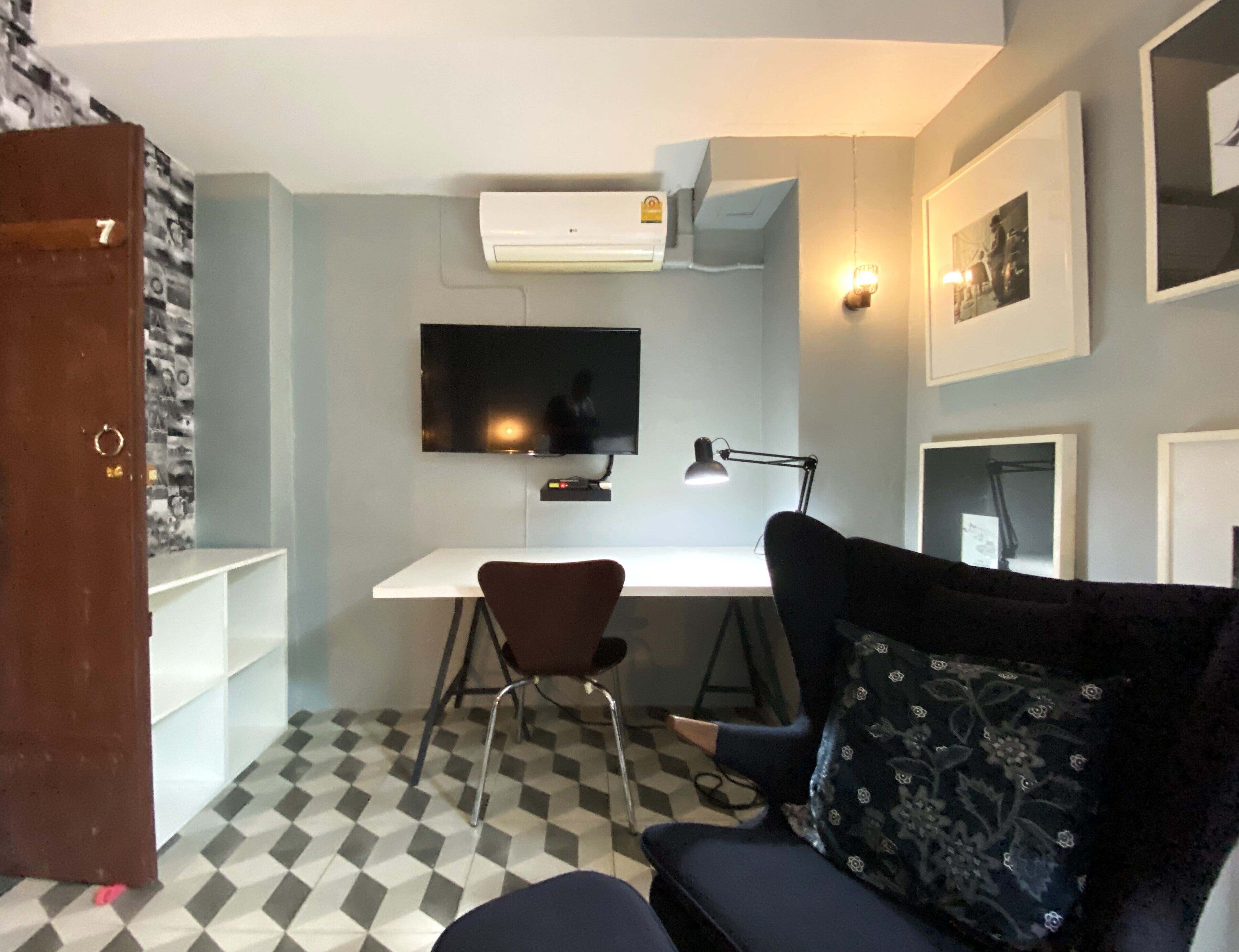 A compact room with a desk, chair, and a sofa. There's a flat-screen TV mounted on the wall, a desk lamp, and a wall-mounted air conditioner. The floor has a checkered pattern.