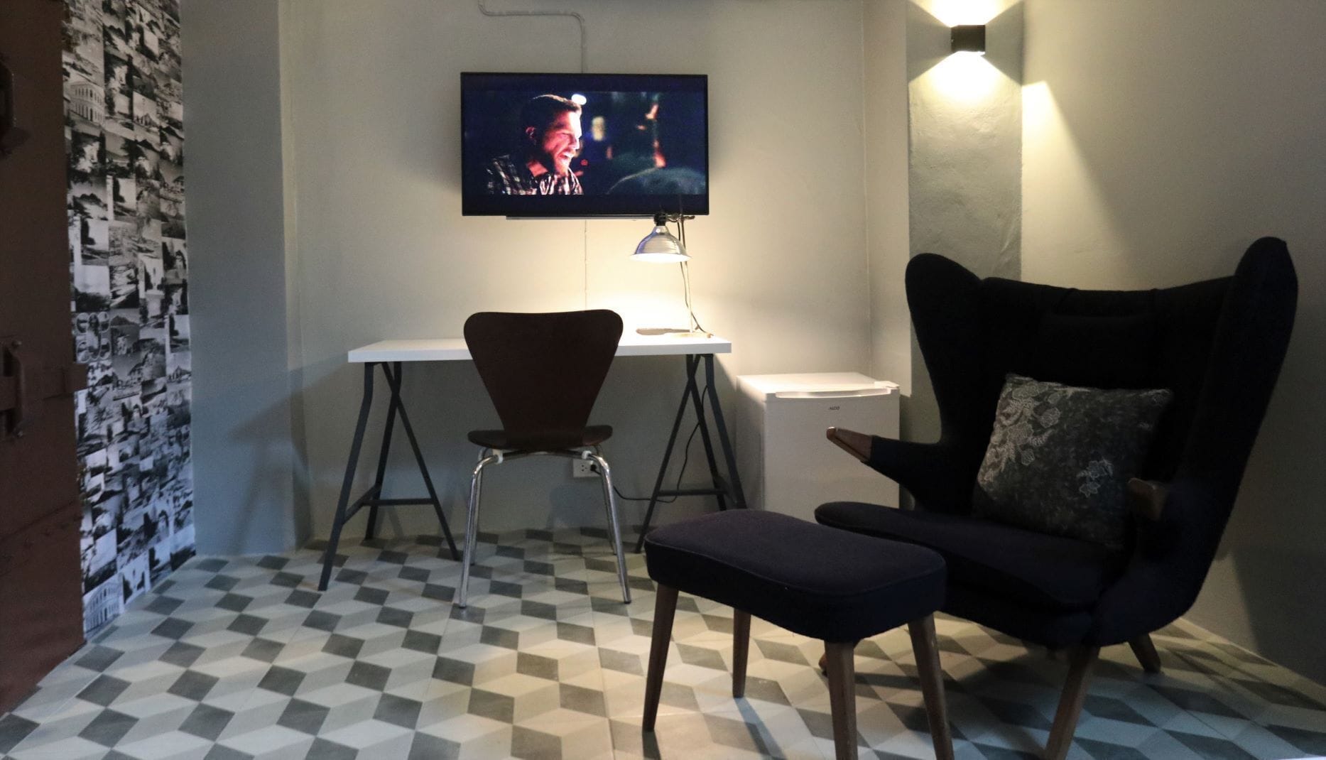 Una habitación con piso a cuadros, un escritorio con una silla, un televisor montado en la pared y una lámpara también fijada a la pared.