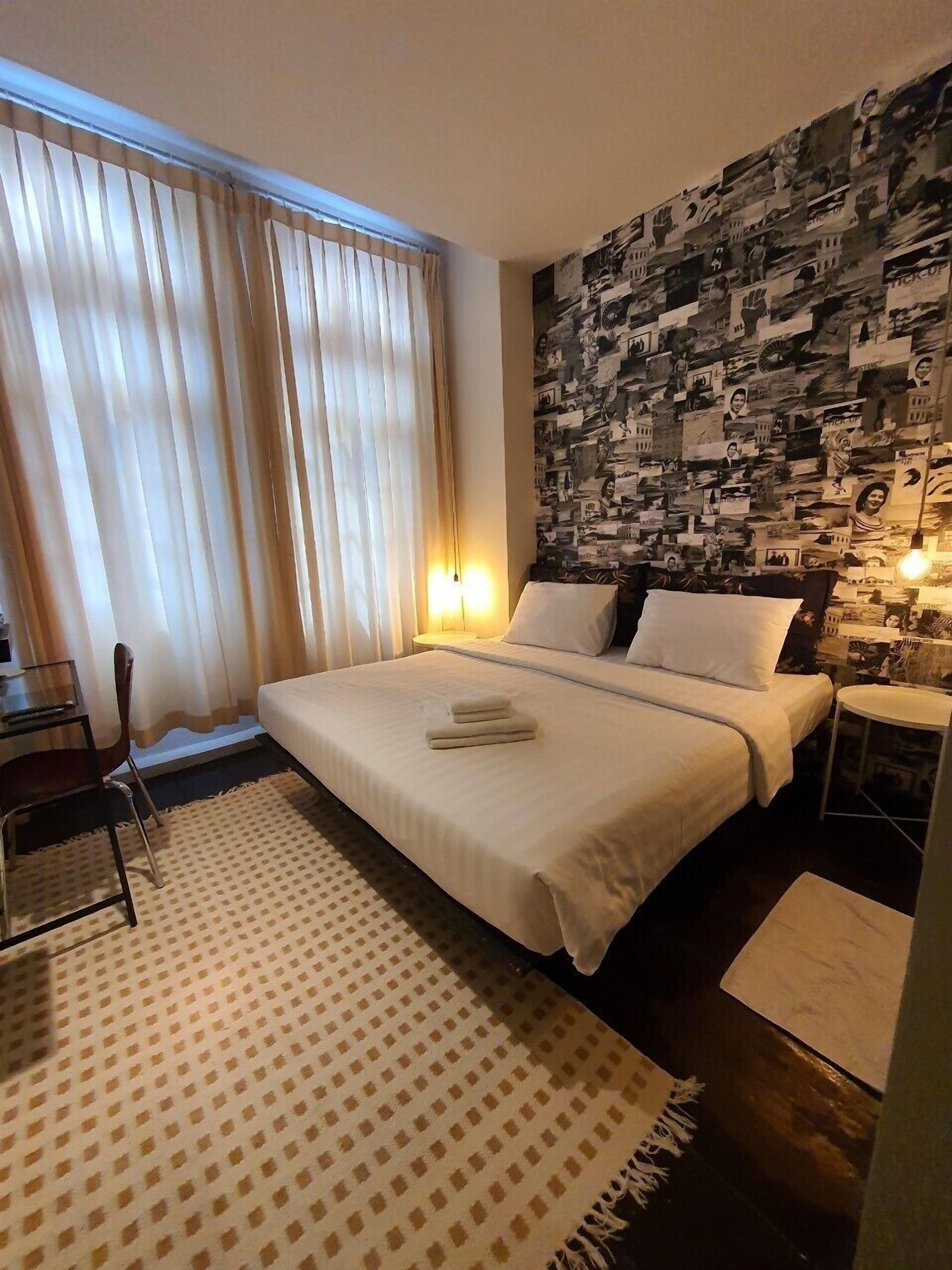 Une chambre d’hôtel avec un lit, un tapis à motifs, un mur orné d’un collage de photos et une fenêtre avec des rideaux.