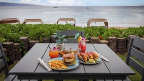 Een tafel gedekt met eten en drinken, met uitzicht op een strand met stoelen en begroeiing.