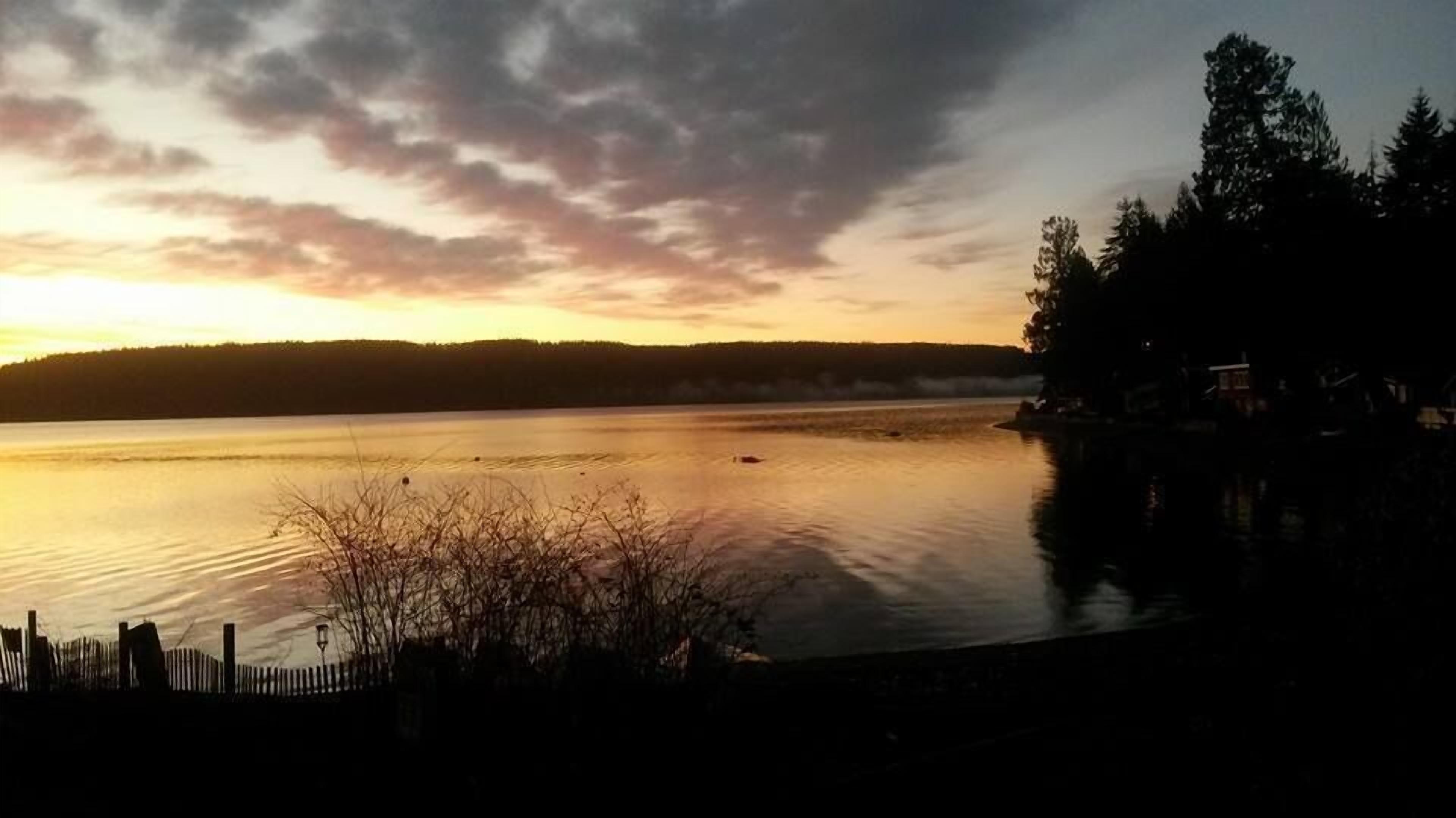 Un lago tranquilo al atardecer con una orilla arbolada y una cerca en primer plano.