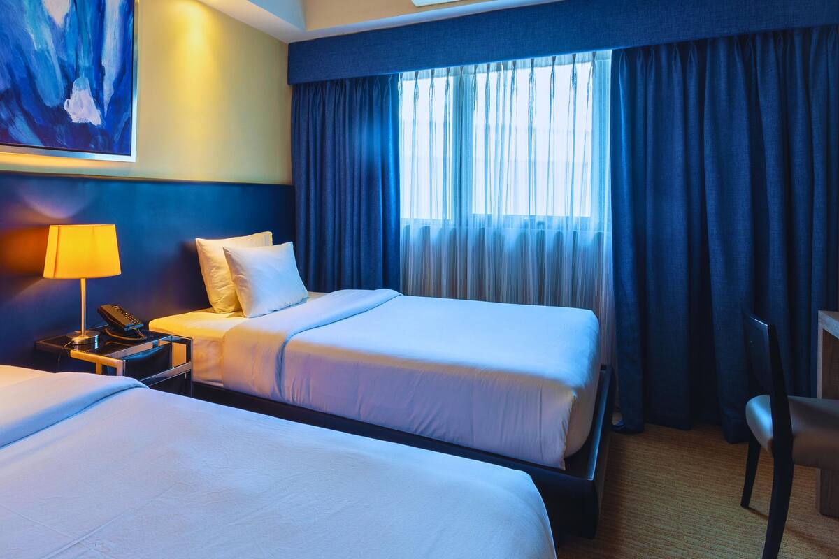 A hotel room with two beds, a nightstand with a lamp, a phone, and a painting on the wall.