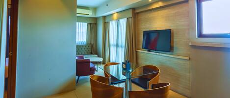 A hotel room with a glass dining table, two wooden chairs, a sofa, and a television mounted on the wall.