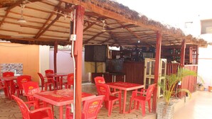 Un espace extérieur abrité, avec des chaises et des tables en plastique rouge, un toit de chaume et un pot de fleurs.