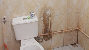 Une salle de bain avec des toilettes, un mur endommagé et une tuyauterie apparente.