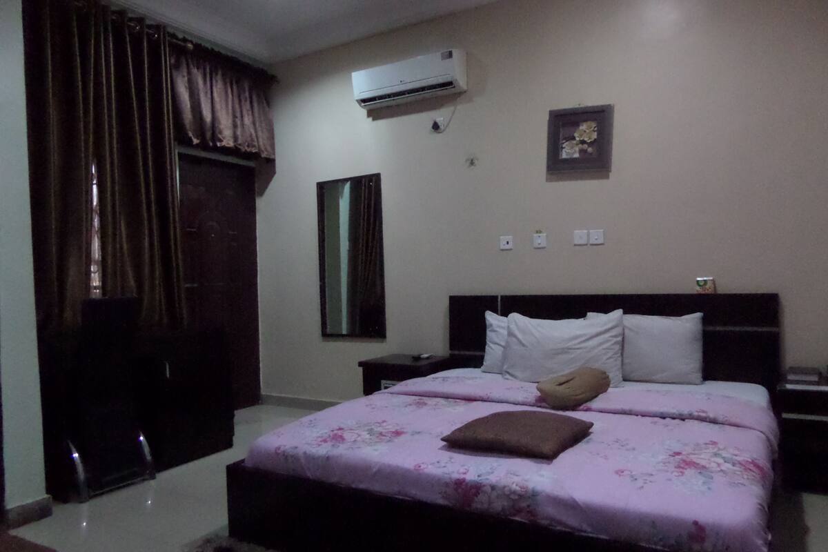 A bedroom with a bed, a nightstand, a dresser, an air conditioning unit, a window with curtains, and a wall-mounted picture.