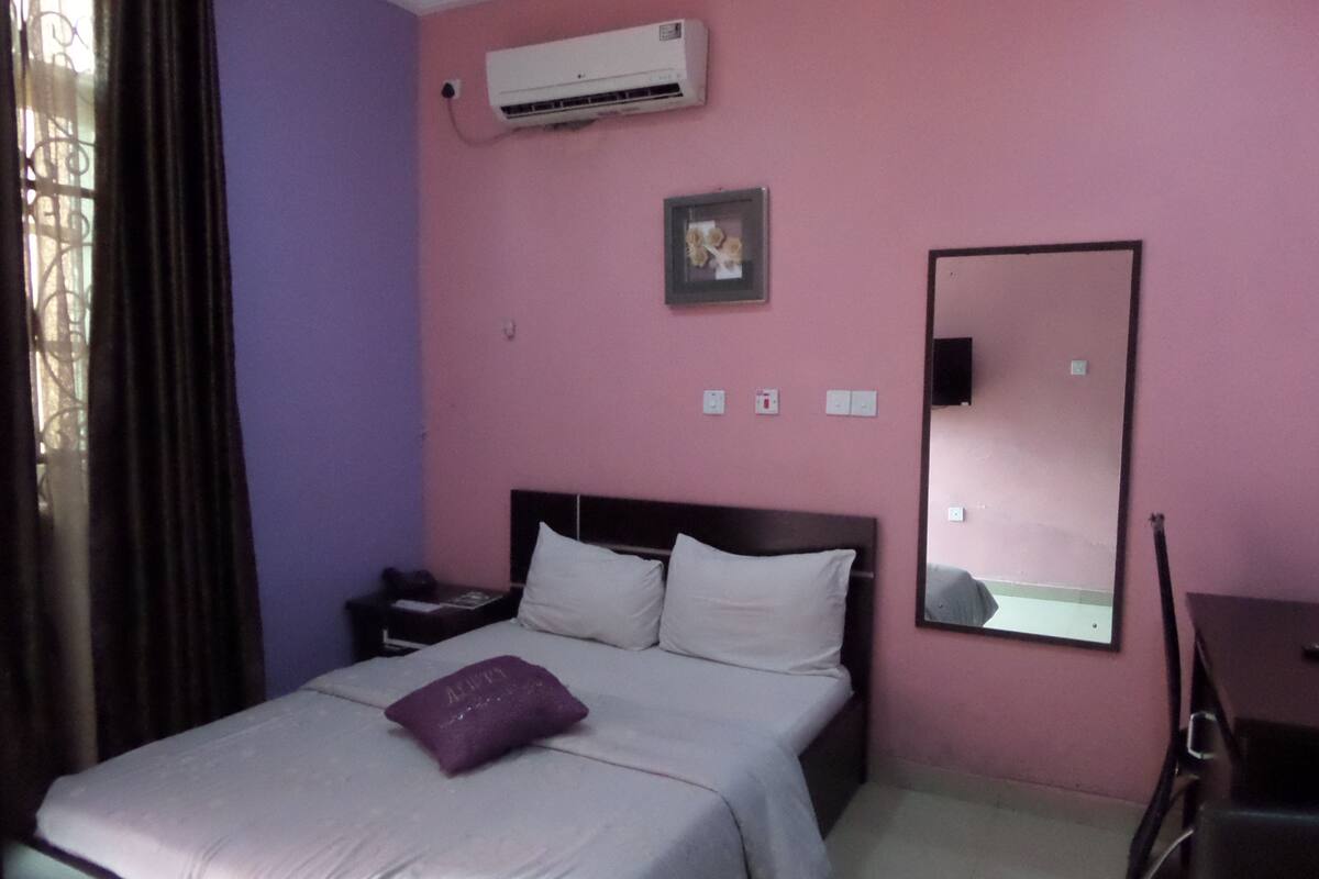 A hotel room with a bed, a nightstand, a desk, a chair, a mirror, and an air conditioning unit on the wall.