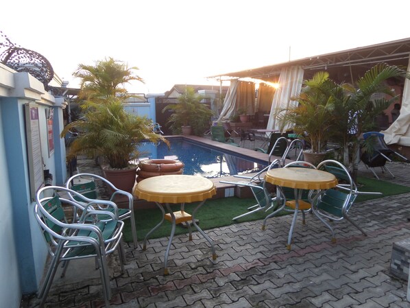 An outdoor pool area with lounge chairs and a table, surrounded by potted plants and a blue building.