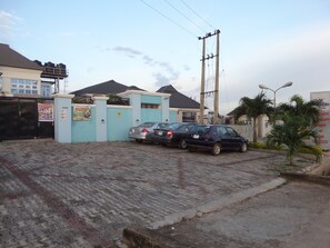A paved driveway with two cars parked, a gated entrance, and a building with a blue facade. - Alheri Suites (Ibadan)