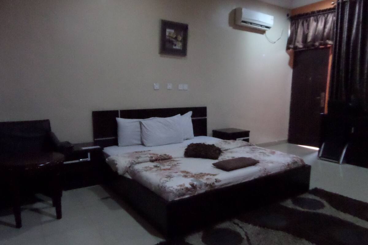 A bedroom with a bed, a nightstand, an air conditioning unit, a chair, and a window with curtains.