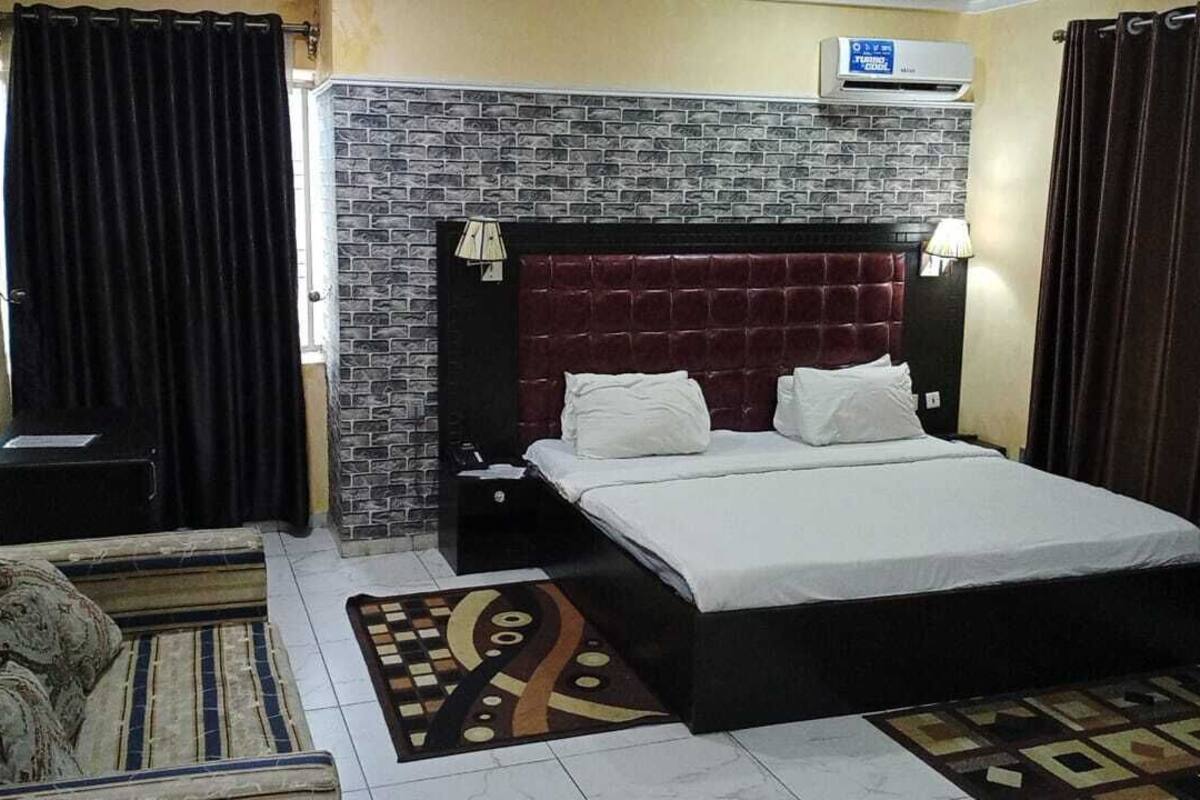 A hotel room with a brick-patterned headboard, a bed with white linens, a nightstand, a lamp, and a decorative rug.