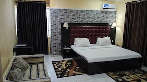 A hotel room with a brick-patterned headboard, a bed with white linens, a nightstand, a lamp, and a decorative rug. - Alheri Suites (Ibadan)
