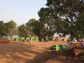 A campsite with multiple green tents, tables, and chairs arranged under trees. - Stay In Pawana Camp (Mawal)