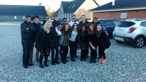 A group of people standing in a gravel parking area with buildings and parked cars in the background.