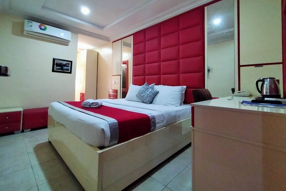 A hotel room with a bed, a bedside table, a mirror, a wardrobe, and an air conditioning unit.
