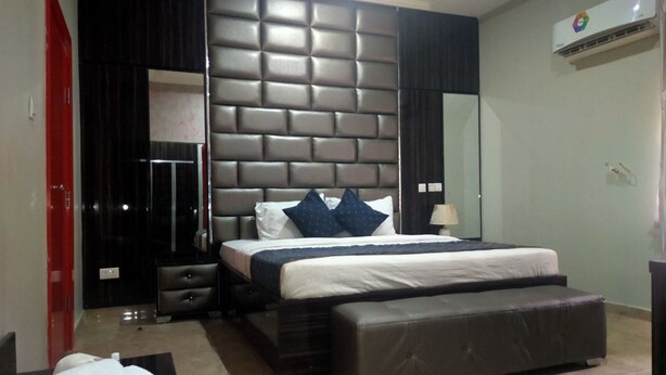 A modern bedroom with a large bed, a padded headboard, a bedside table, a lamp, and a red door. - BoardWalk Hotel Wuse 2 (Abuja)