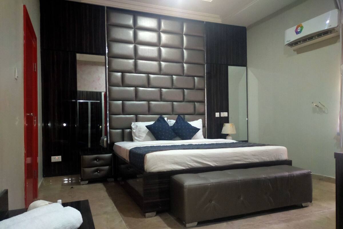 A modern bedroom with a large bed, a padded headboard, a bedside table, a lamp, and a red door.
