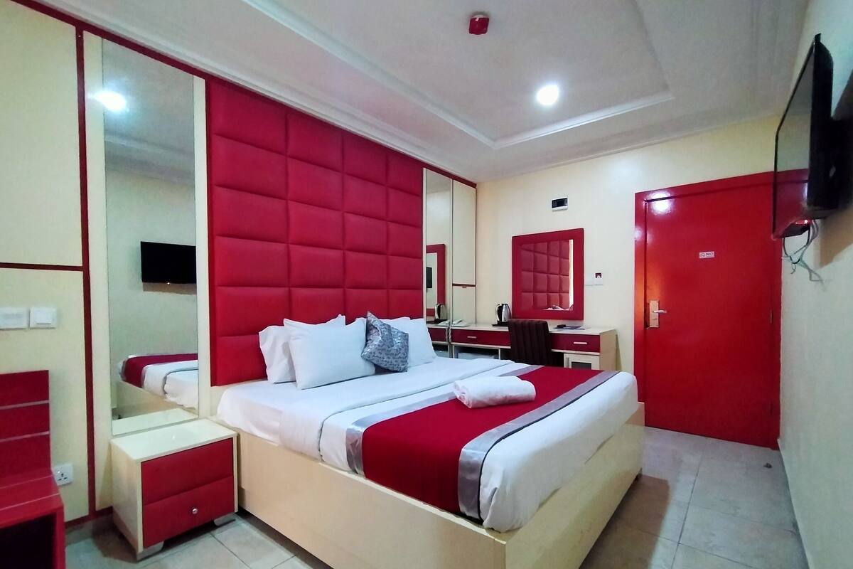 A modern hotel room with a large bed, a desk, a chair, a wardrobe, and a TV.