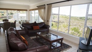 A spacious living room with leather sofas, a glass coffee table, and large windows offering a view of the countryside. - 2 Sheerline (Knysna)