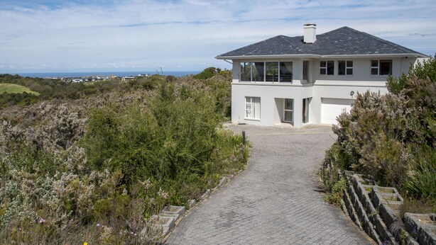 A two-story house with a paved driveway leading to the entrance, surrounded by dense shrubbery and a clear sky. - 2 Sheerline (Knysna)