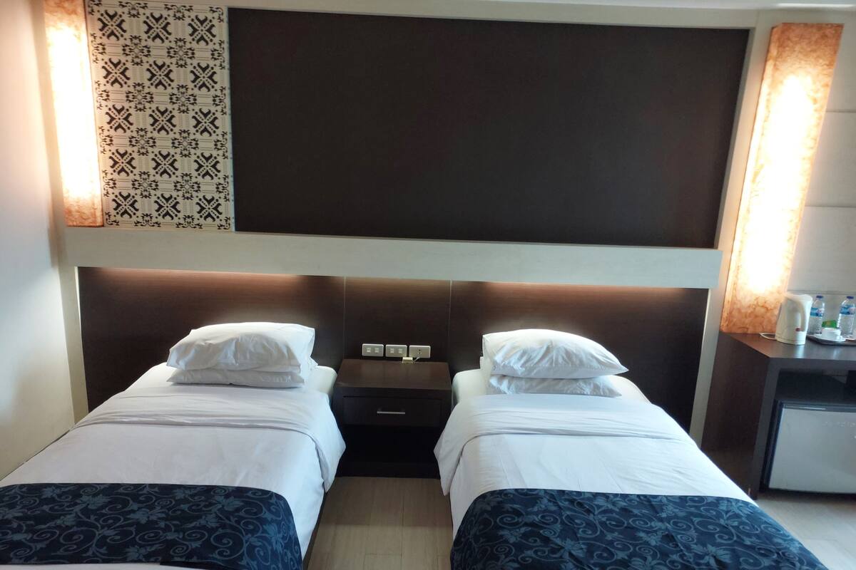 A hotel room with two single beds, a bedside table with a lamp, a small desk with a chair, and a patterned wall decoration.