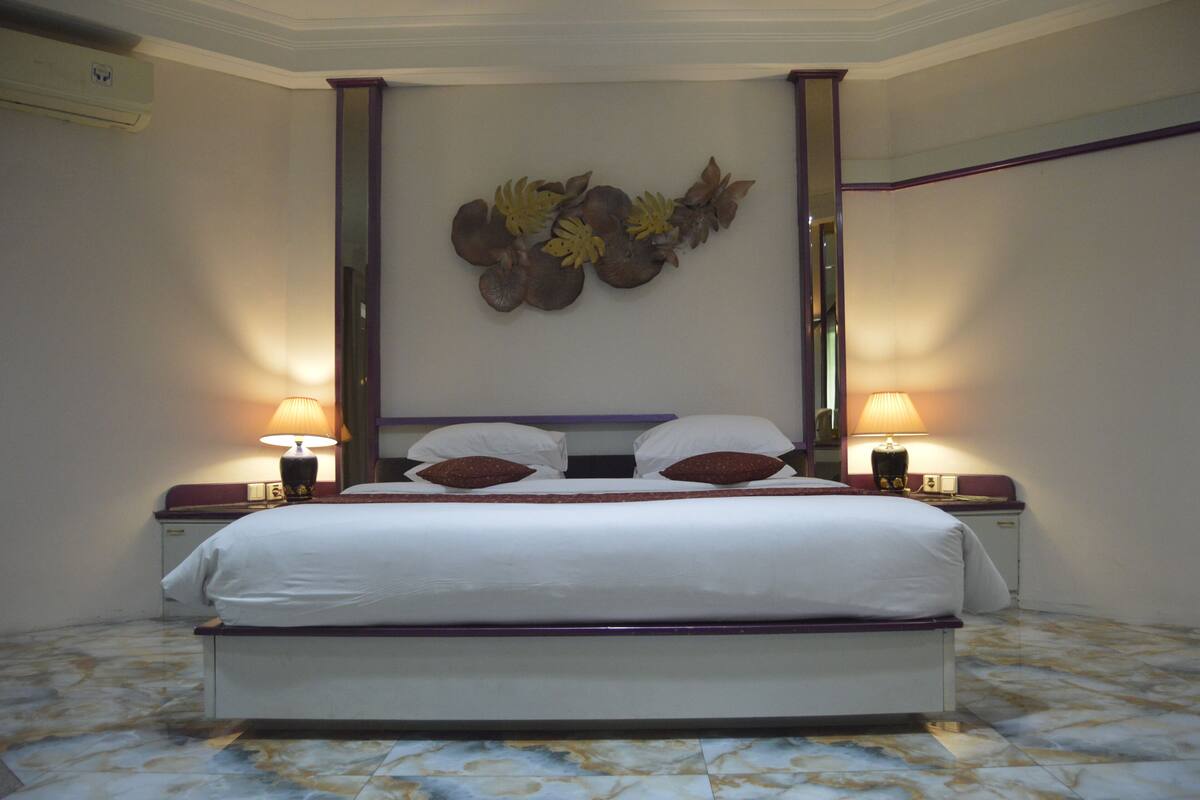 A neatly made bed with white linens and two decorative pillows, flanked by two bedside tables with lamps, in a room with a marble floor and a wall-mounted decorative piece above the bed.