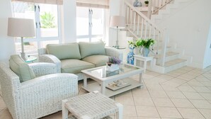 A bright living room with a beige sofa, wicker chairs, a glass coffee table, and a staircase with white railings. - 8 Sheerline (Knysna)