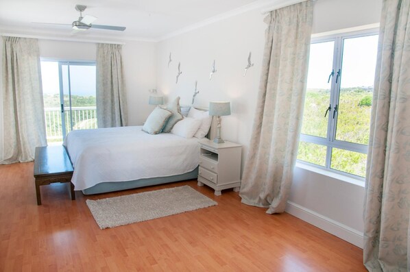 A bedroom with a bed, bedside tables, a ceiling fan, and a view of greenery through the window.