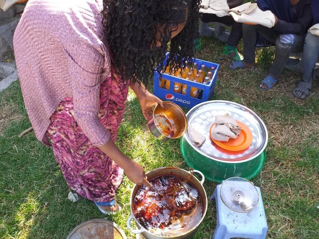 A person cooking outdoors with a pot on a portable stove, a large green container, and a blue crate of Pepsi bottles.